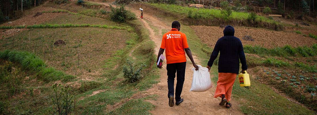 a oncology program coordinator and social worker walk to a patient’s home in Burera District, Rwanda. They are carrying a food support package.
