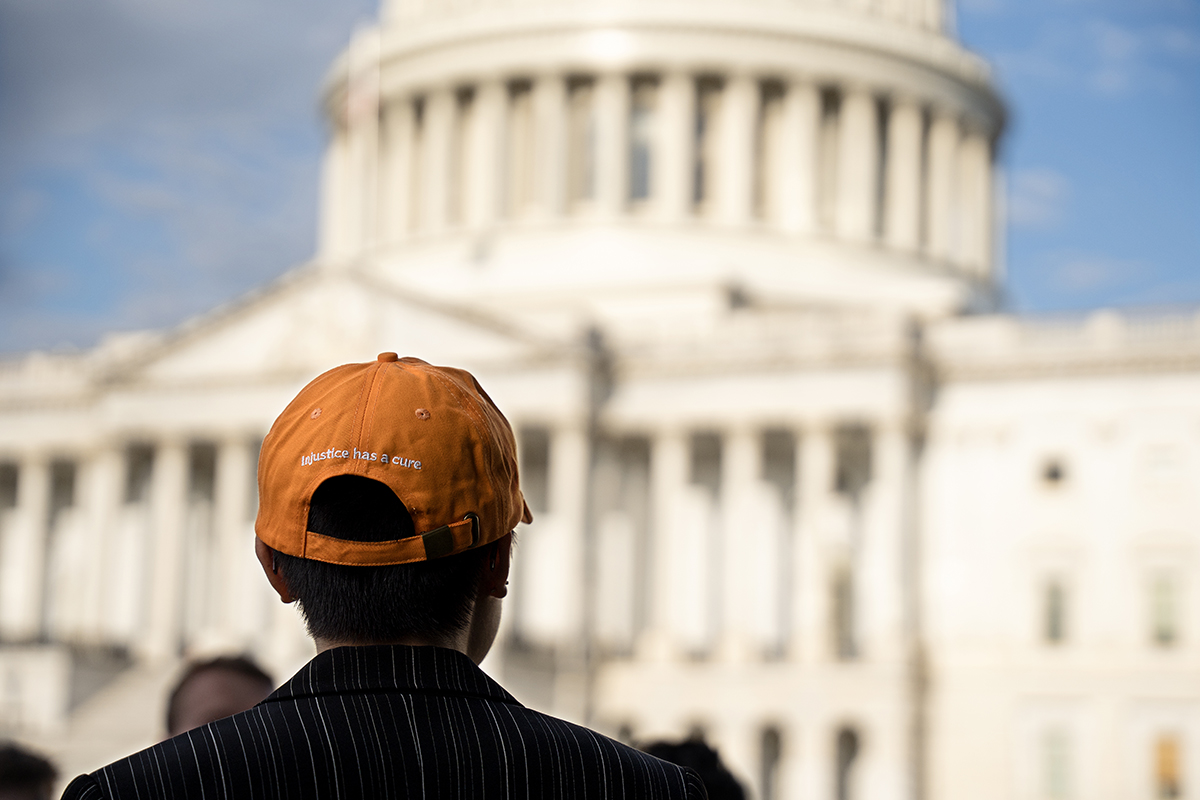 Person in an orange hat faces away from the camera with the U.S. Capitol building blurry in the background. The back of the hat says "Injustice has a cure."