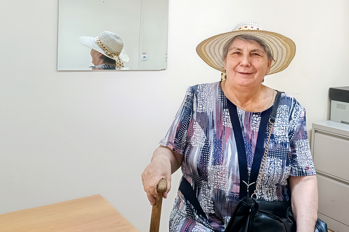 a woman wearing a wide brimmed hat smiles at the camera wearing a purple patterned shirt while holding a cane