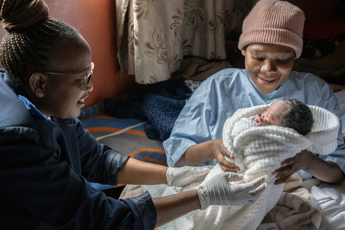 a nurse smiling and wearing gloves hands a newborn wrapped in a white blanket to its mother wearing a blue hospital gown and pink hat who is smiling at her