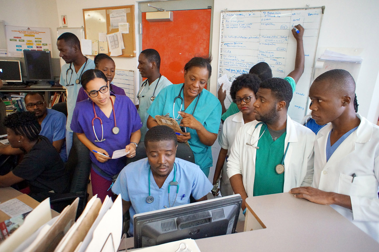 Morning shift change in the emergency department at PIH-supported University Hospital in Mirebalais, Haiti. They have just finished visiting each patient's bedside and the night shift is passing info along to the morning shift physicians. The white board is being updated with patient names and statuses. Residents are also going over patient files in the electronic medical records system. The varying colored scrubs indicate which year of residency the emergency medicine and internal medicine doctors are in.