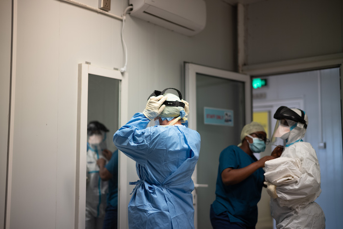 Caregiver in hazmat suit putting on their mask while another person watches them put