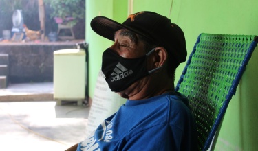 elderly patient sits on his front porch at home in Chiapas, Mexico. He wears a face mask.