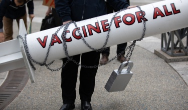 an oversized, white prop vaccine with gray chains and a lock around it that say "VACCINE FOR ALL" in red 