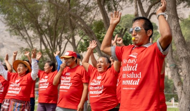 Outside in front of trees, people in red shirts that say "No hay salud sin salud mental" raise their arms in the air, the person closest to the camera has a whistle in their mouth