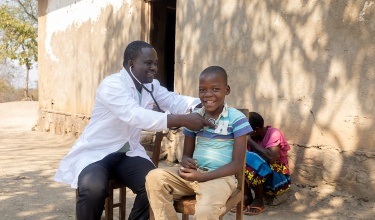 Clinical team member Henderson Masanjala holds a stethoscope to the chest of a smiling Promise Douglas.