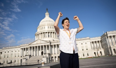 Fynn standing in front of the U.S. Capitol building with their arms raised triumphantly under a bright blue sky