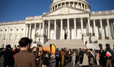 A large group of people gathered at the steps to the capitol being guided by a person holding an orange megaphone