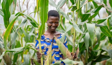 Uwizeyimana Garas stands amidst tall maize, smiling in a blue dress