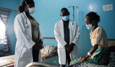 A patient wearing a mask sits on a bed while two clinicians in masks and white coats stand beside her with their arms clasped in front of their waists
