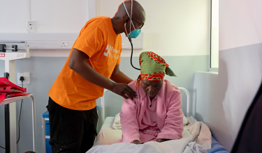 A person in an orange PIH shirt with a mask on holds a stethoscope to the back of a woman wearing pink sitting hunched over in a hospital bed.