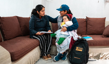 a person with a blue Socios En Salud hat holds a child wrapped in blankets and puts her hand on the shoulder of a woman to her right holding papers