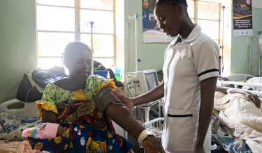 a nurse stands beside a partient sitting with a baby in her lap with a blood pressure cuff around her upper arm