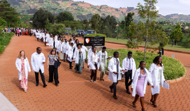 A long line of students in white coats and formal attire walk along a road.