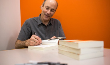 Tracy Kidder sits at a round table against an orange wall signing books with a sharpie with a smile on his face
