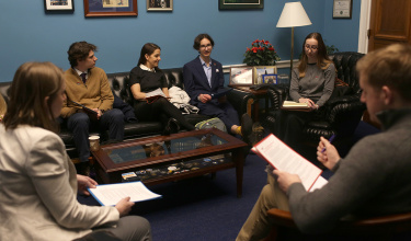 In a room with blue walls, several people sit on a leather couch and chairs for a meeting