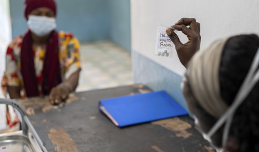 Medication in a small bag is being handed to a patient who sits across a table